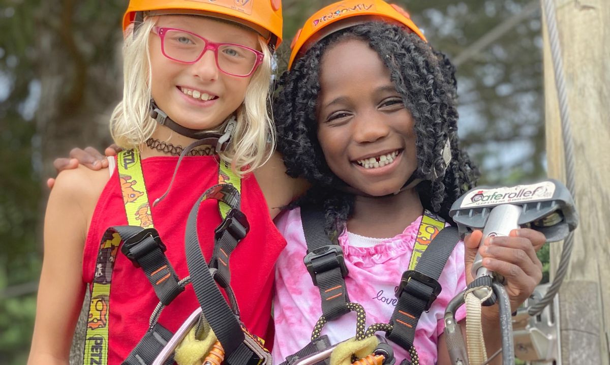 Two smiling young girls wearing safety harnesses and helmets enjoy a ziplining adventure in Hocking Hills, Ohio, during a fun outdoor family trip.