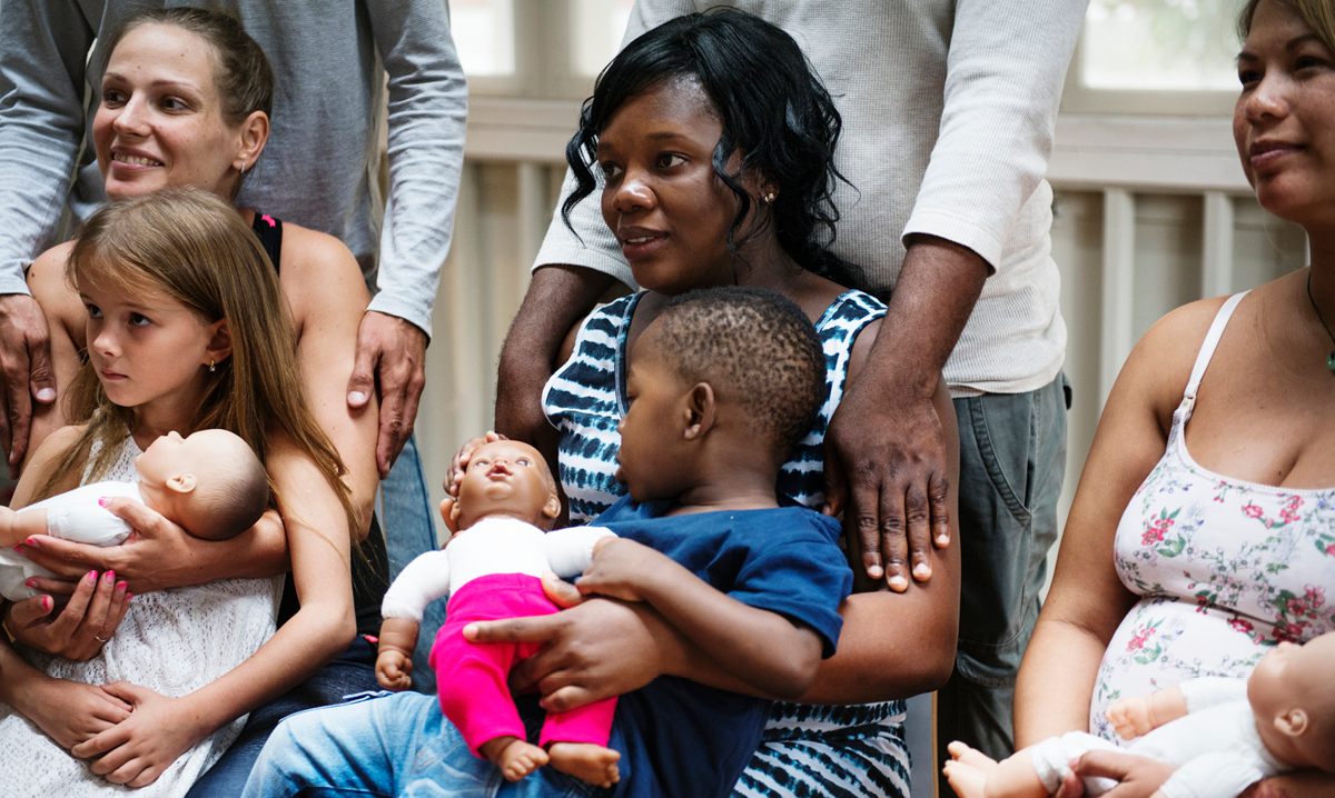 Three moms with toddlers sitting in a char with people standing behind them