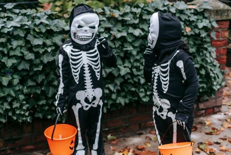 Two kids dressed in skeleton costumes hold orange buckets while trick-or-treating during a family-friendly Halloween event outdoors.