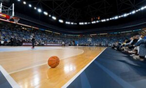 A basketball sits on the court at a packed arena, with fans watching and players warming up before the game.