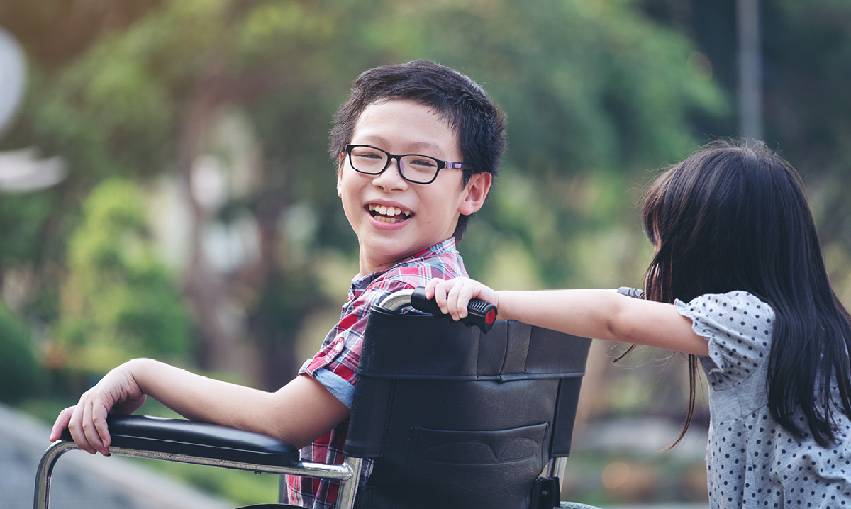 A smiling boy in a wheelchair is pushed by a young girl outdoors, representing inclusive special needs events for kids in Detroit.