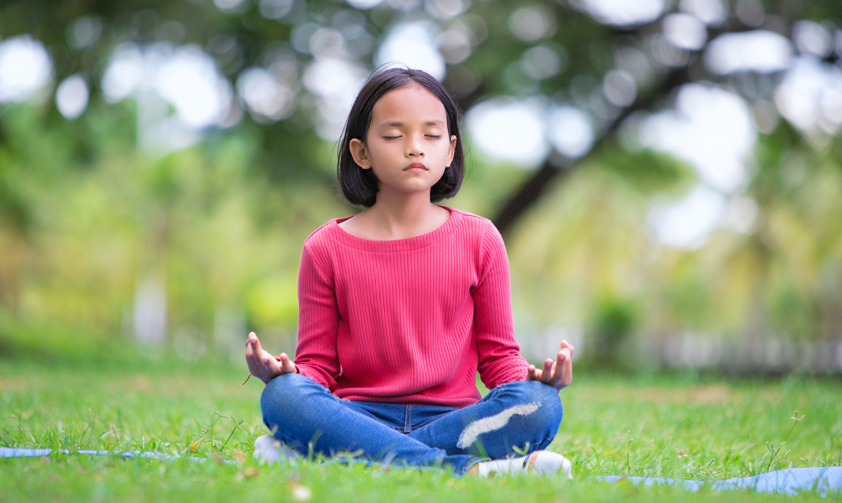 Little girl in pink shirt sitting in the grass while meditating with her eyes closed a