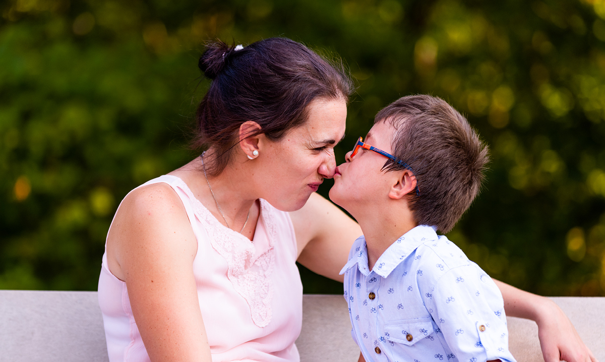 A boy with red glasses kisses mom's nose on a bench