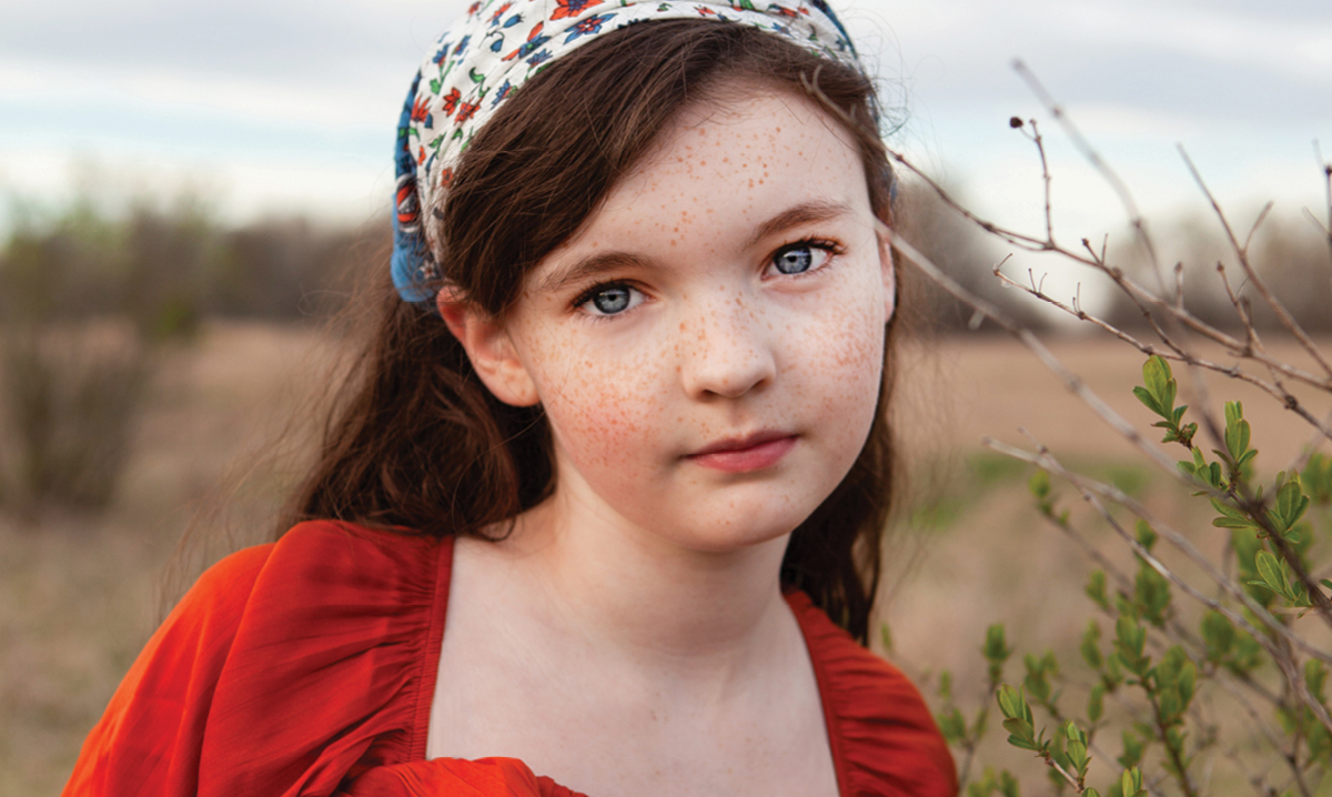 Sydney is wearing a red dress and bonnet while standing in a field and looking at the camera