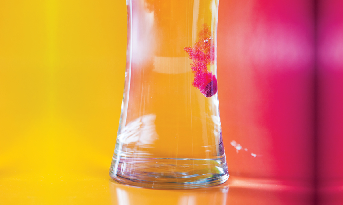 Glass of water on a yellow and pink background with a Crayola Color Bath Drop fizzing in it