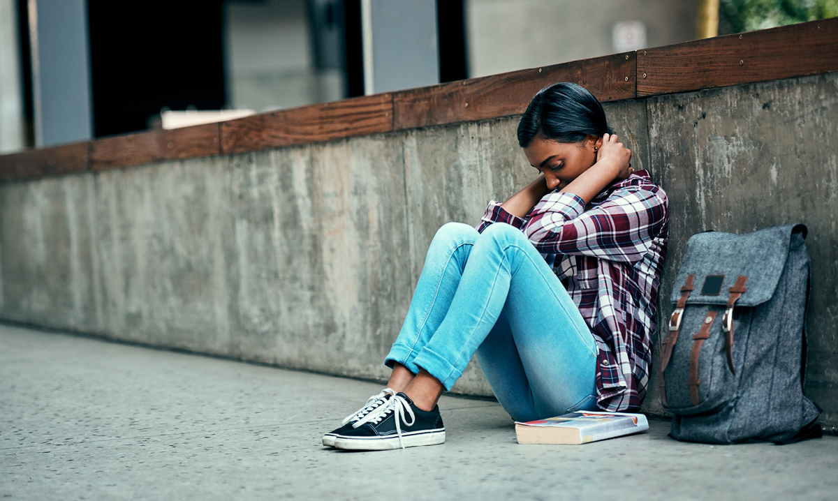 Girl puts her head in her hands while sitting on the ground next to a book bag