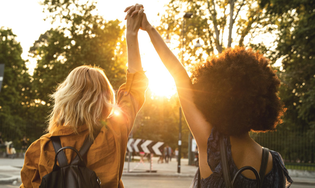 A white woman and a black woman holding lifted hands as they walk toward a sunrise