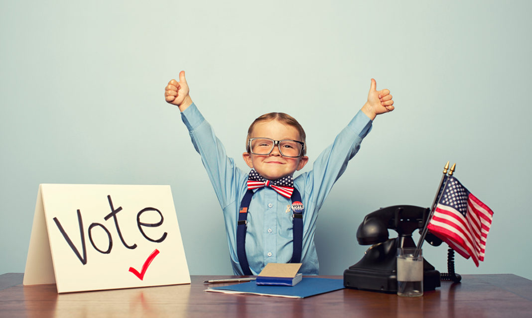 Kid in a bow tie puts thumbs up in the air behind a desk