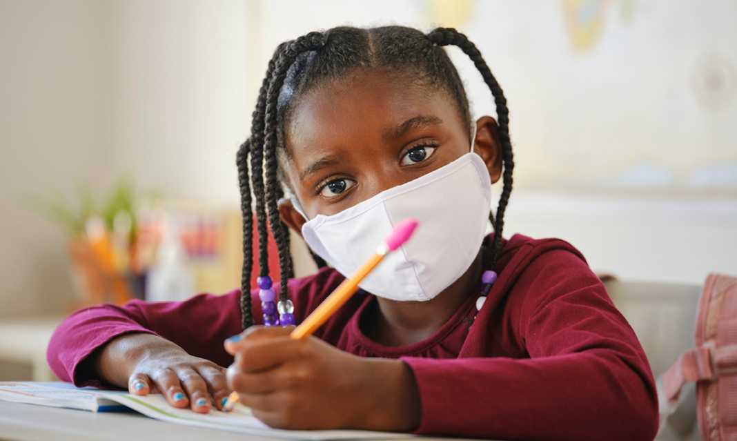 Young girl wearing a mask holding a pencil and looking at the camera