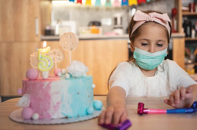 Girl in a mask reaching for a party favor while sitting next to a birthday cake