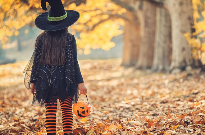 The back of a child, dressed as a witch, walking among yellow and orange trees.
