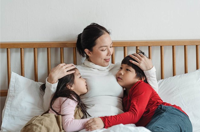 An asian woman hugging two young children who are looking at her while sitting on the couch