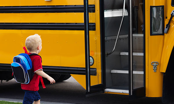 Kindergartner with a red shirt and blue backpack getting on a school bus