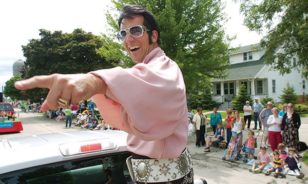 Elvis tribute artists in a pink shirt points while sitting on a car