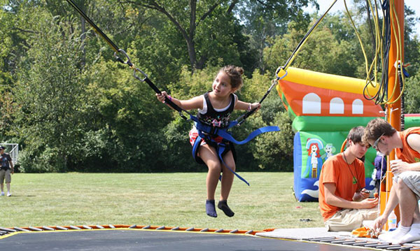 Young girl bounces on a trampoline with harnesses at the Franklin Labor Day Round Up, with inflatable games and kids in the background.