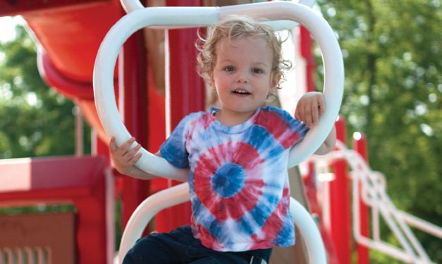 A young child in a colorful tie-dye shirt enjoys playing on a red playground structure on a sunny day. The outdoor setting with greenery in the background provides a fun and vibrant atmosphere.