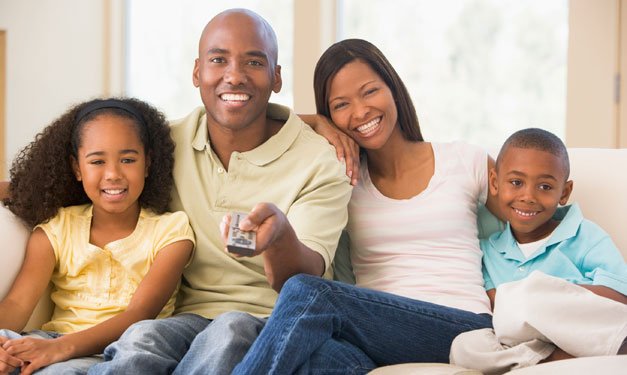 A family sitting on a couch as dad points the remote toward a tv out of shot
