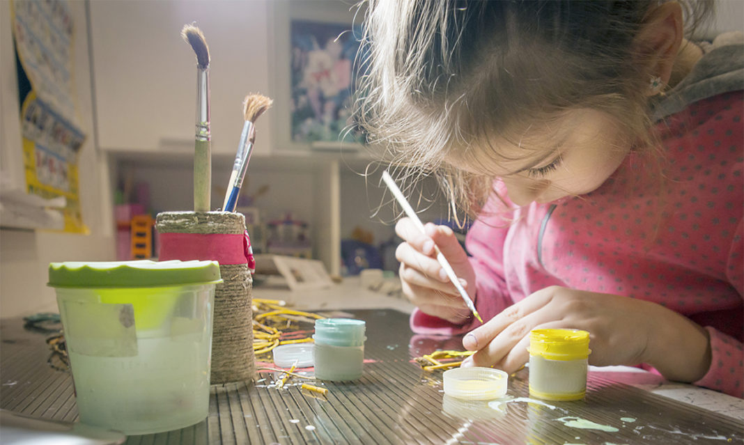 A young child focuses on painting a craft project at home, surrounded by art supplies including brushes and small paint containers.