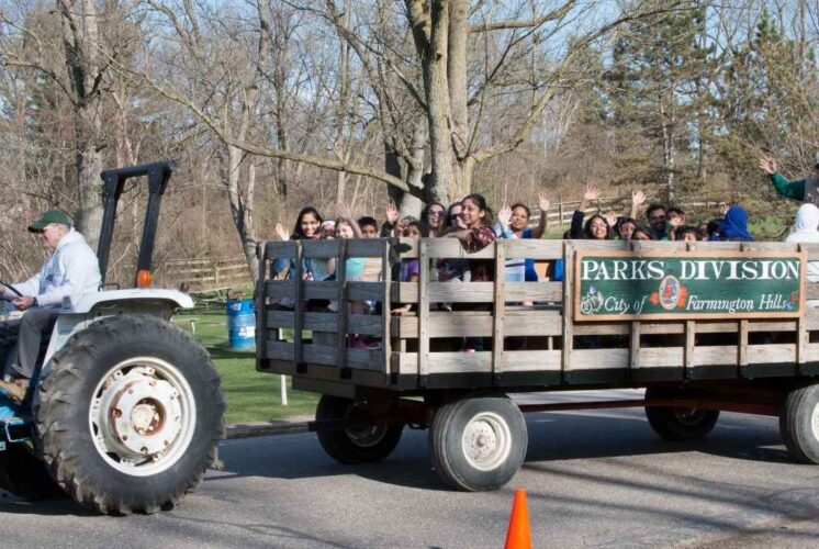 Families enjoy a hayride at Heritage Park during Hay Day in Farmington Hills with kids and parents waving while riding a tractor-pulled wagon.
