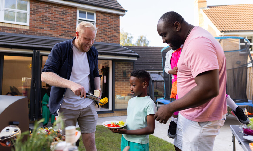 Two dads and a child enjoy an autism-friendly backyard BBQ, sharing food and laughter in a relaxed, supportive environment.