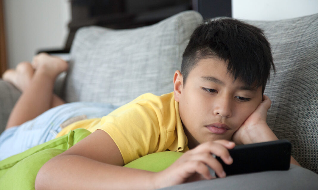A young boy in a yellow shirt lying on a couch, intently looking at a smartphone, illustrating screen time for kids in a relaxed home setting.