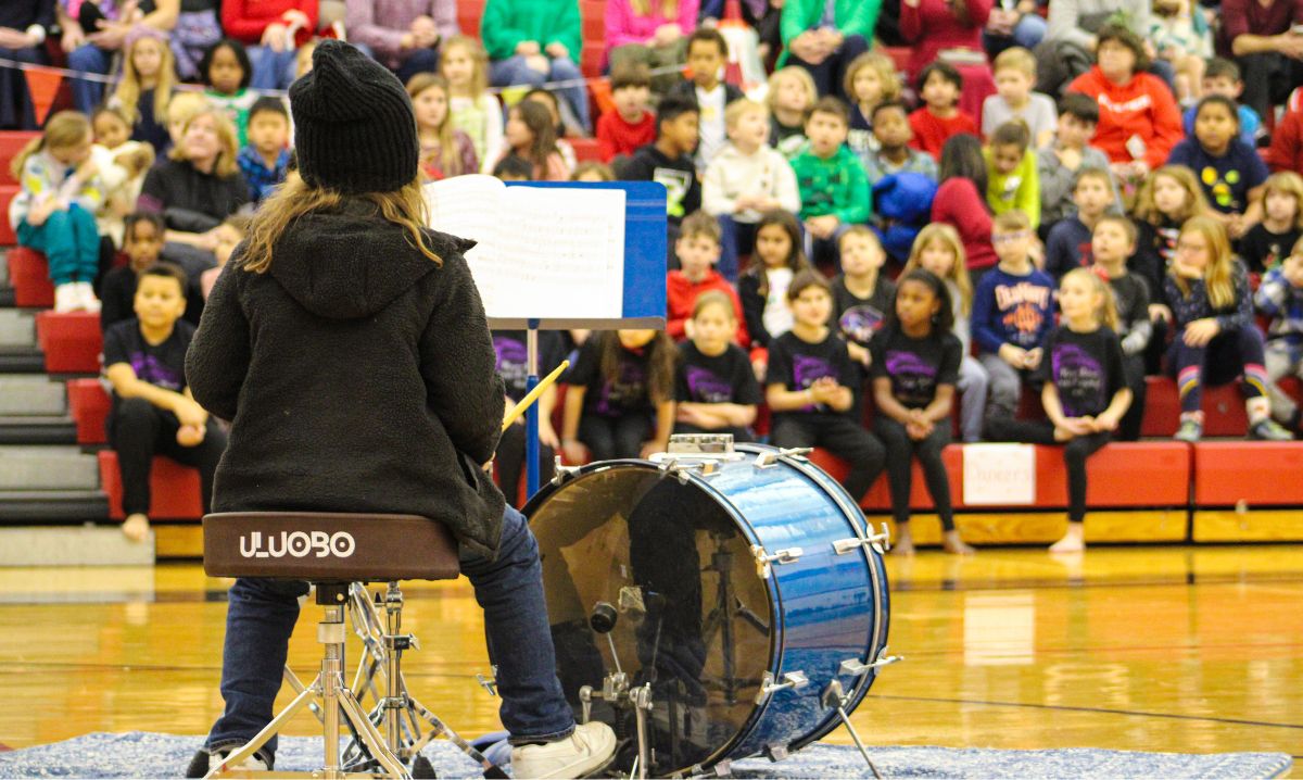 A student at The Roeper School performs on the drums during a school assembly with classmates watching in the background.