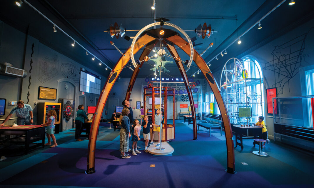 A family explores a colorful and interactive science exhibit at a hands-on museum. The room features unique structures, educational displays, and engaging activities designed to inspire curiosity and learning.