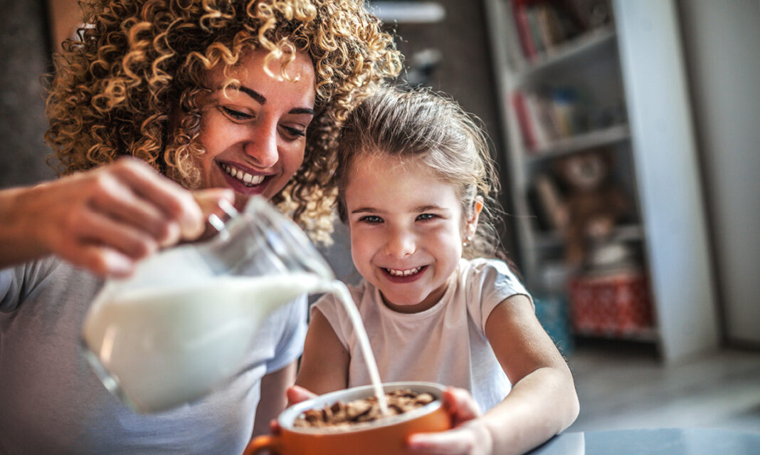 A smiling mother pours milk into her daughter's cereal bowl as they enjoy breakfast together in a cozy home kitchen.