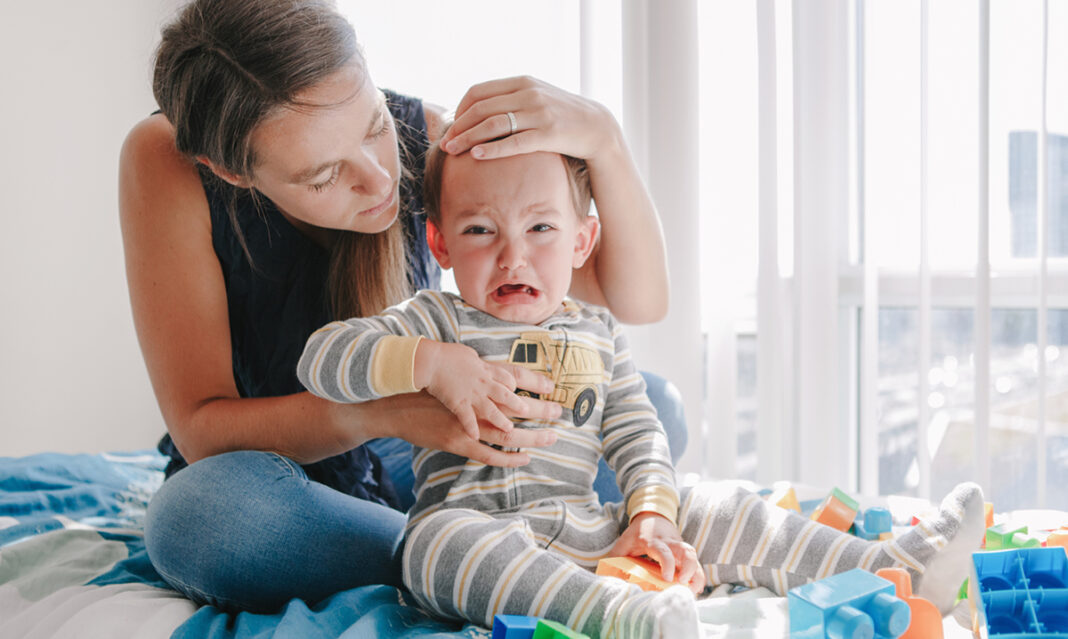 A crying baby sitting on a bed with toys as their mom gently holds their head after headbutting — related to toddler headbutting behavior and care