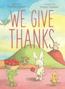 Cover of the children's book We Give Thanks by Cynthia Rylant, illustrated by Sergio Ruzzier. The whimsical illustration features a bunny, a frog carrying a basket of apples, and other cheerful animals in a scenic outdoor setting, emphasizing themes of gratitude and friendship.