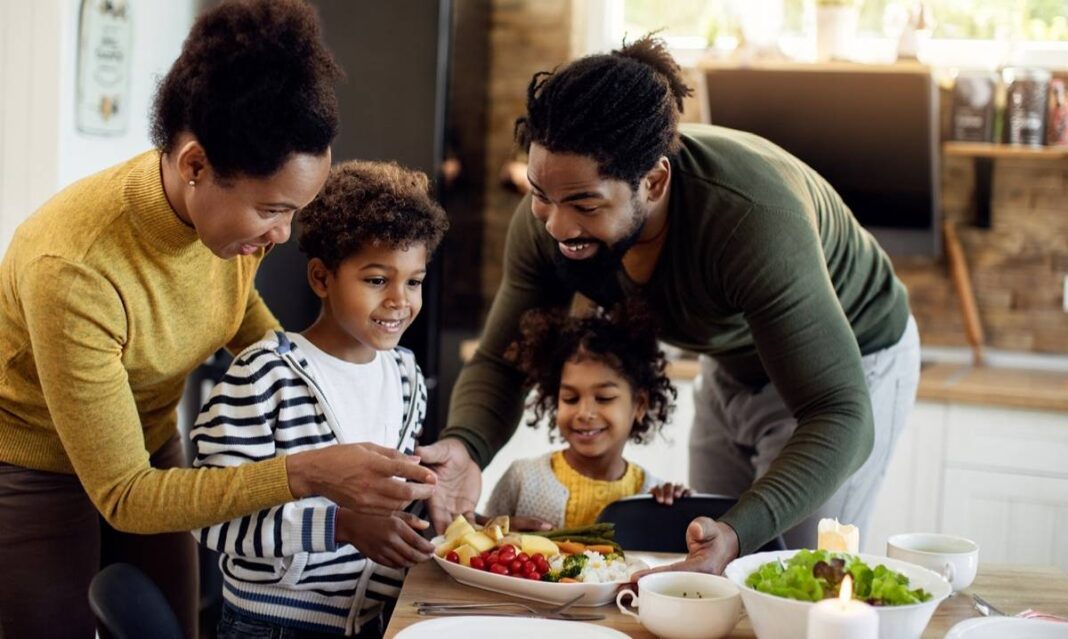 Parents and young children gathered around a Thanksgiving table with vegetables and healthy dishes, smiling and enjoying a relaxed family meal.