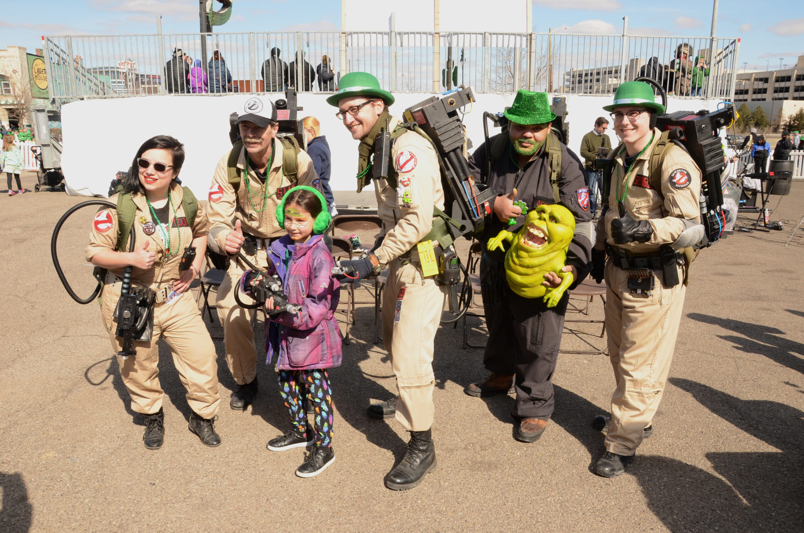 A group of people dressed as Ghostbusters pose with a child at a St. Patrick’s Day parade, wearing festive green hats and carrying proton packs. One participant holds a Slimer prop, adding to the fun atmosphere of the event.