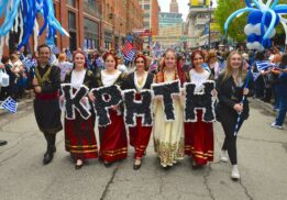 Participants in traditional Greek costumes holding signs and flags during the Detroit Greek Independence Parade 2025, celebrating Greek culture and heritage.