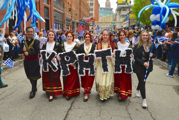 Participants in traditional Greek costumes holding signs and flags during the Detroit Greek Independence Parade 2025, celebrating Greek culture and heritage.