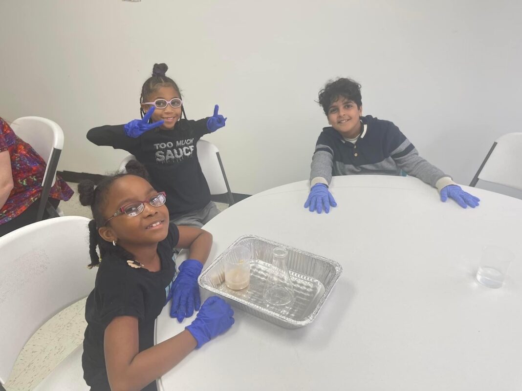 Three elementary school students smiling and wearing gloves during a hands-on STEM activity, representing the benefits of flexible learning in a creative classroom environment.
