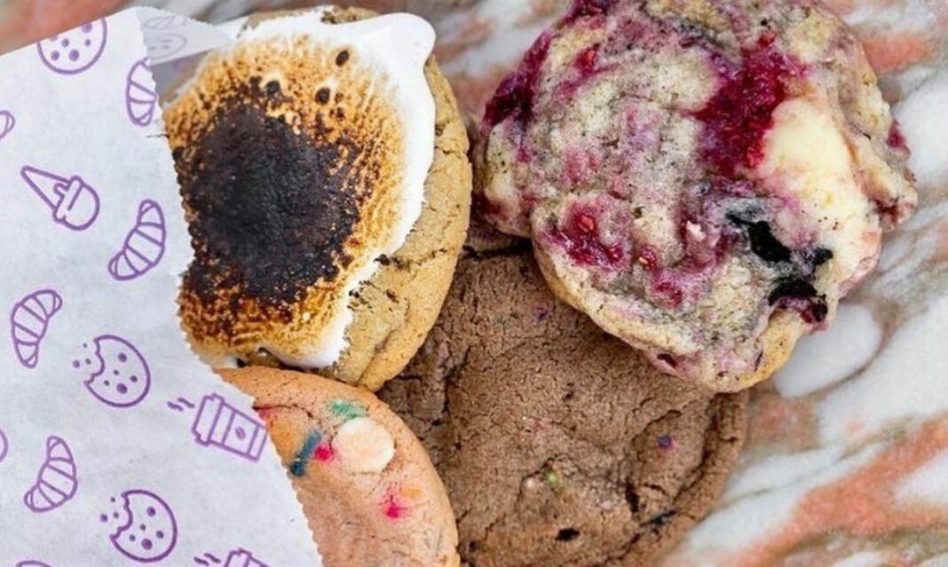 A selection of gourmet cookies displayed on a marble background, including a toasted marshmallow cookie, a berry-filled cookie, a chocolate cookie, and a colorful funfetti cookie wrapped in decorative paper. Perfect for dessert lovers.
