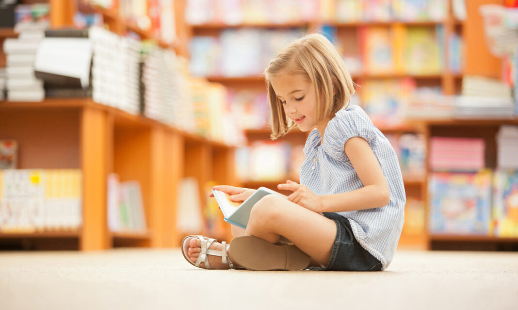 A young girl sitting on the floor in a library, engrossed in a book, showcasing efforts to foster a love of reading in children.