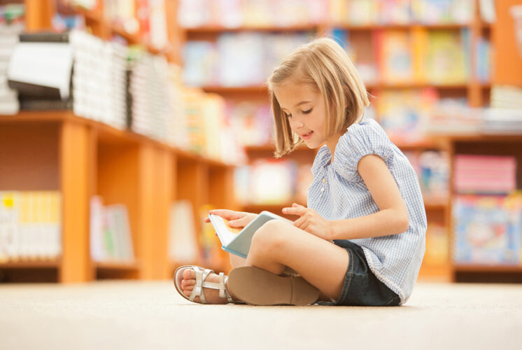 A young girl sitting on the floor in a library, engrossed in a book, showcasing efforts to foster a love of reading in children.