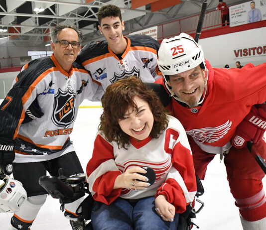MI-UCP Skating to Close the Disability Divide Charity Hockey Event at St. Mary’s Arena A smiling hockey fan in a wheelchair poses on the ice with Detroit Red Wings player Darren McCarty and other hockey players at a charity event. The inclusive event brings together athletes and fans for a special community experience.