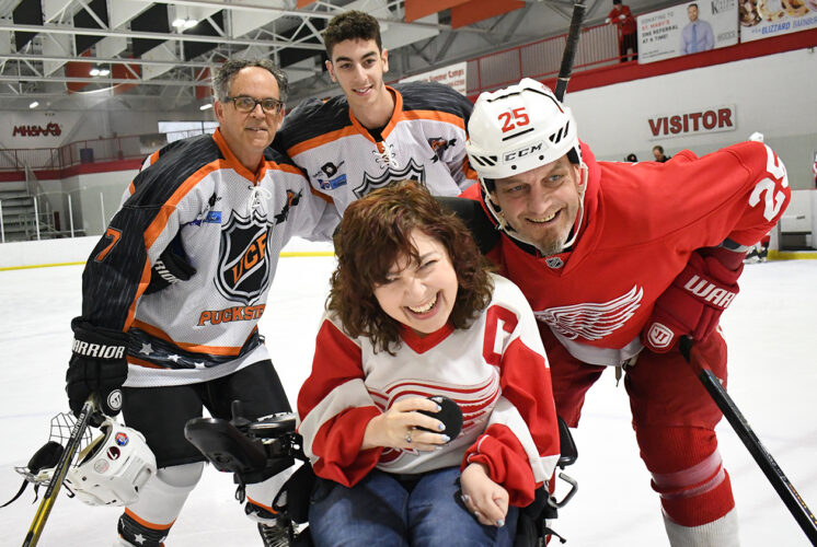 A smiling hockey fan in a wheelchair poses on the ice with Detroit Red Wings player Darren McCarty and other hockey players at a charity event. The inclusive event brings together athletes and fans for a special community experience.