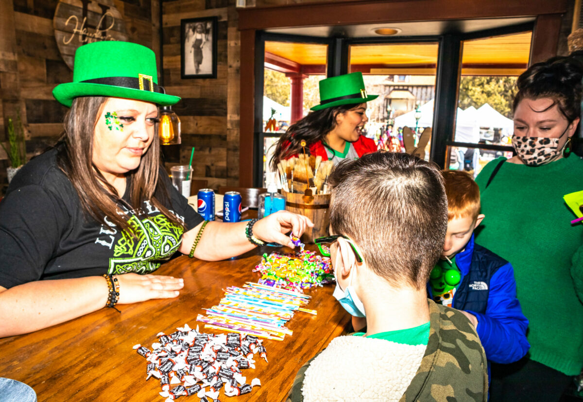 A festive St. Patrick’s Day event where children receive candy from a booth attendant wearing a green hat and shamrock face decoration. Families dressed in green celebrate indoors, enjoying holiday-themed activities and treats.