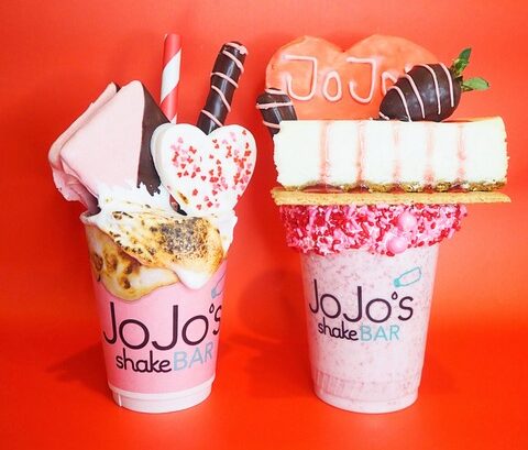 Two extravagant milkshakes from JoJo's Shake Bar, featuring over-the-top toppings like cheesecake slices, chocolate-dipped strawberries, marshmallows, and heart-shaped cookies, set against a vibrant red background. Perfect for dessert enthusiasts.