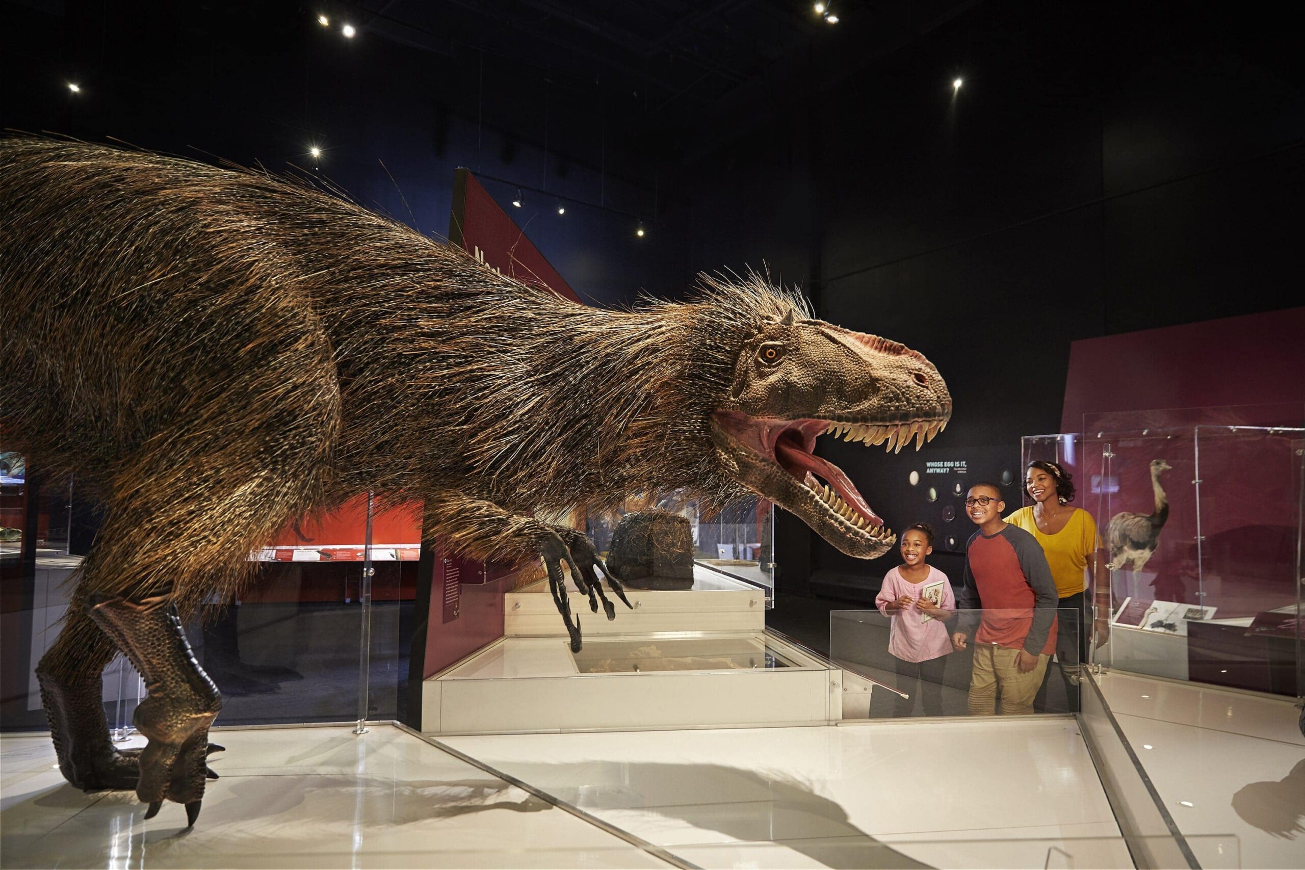 Family smiles while viewing a lifelike feathered dinosaur exhibit at the COSI Center of Science and Industry’s Dinosaur Gallery in Ohio.