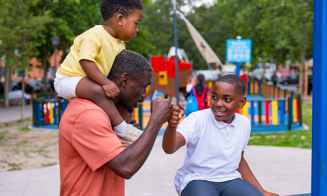 African black ethnicity father waving with his children in the playground of the city park