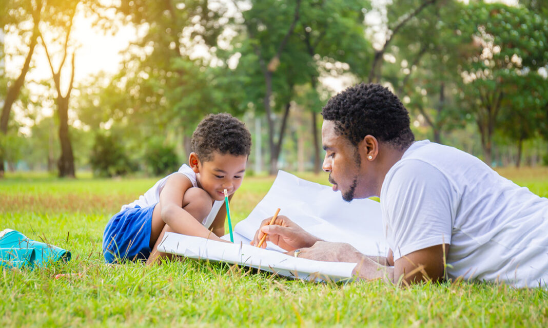 A Black father and his young son enjoy a summer learning activity together in a metro Detroit park, drawing in a large sketchbook while lying on the grass surrounded by trees and sunlight.