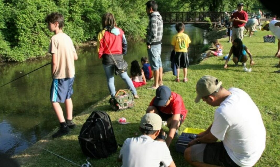 Families and kids fishing by a small creek on a sunny day, with fishing gear and backpacks spread out on the grassy bank.