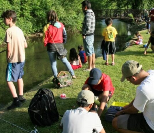 Best Things to Do with Metro Detroit Kids in September Families and kids fishing by a small creek on a sunny day, with fishing gear and backpacks spread out on the grassy bank.