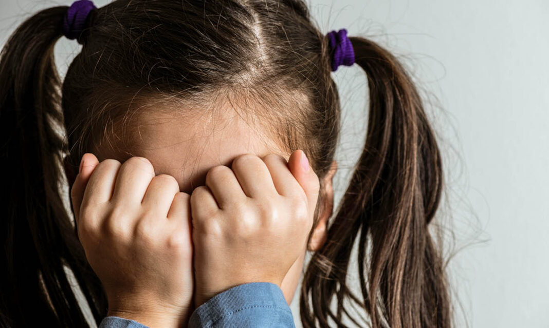A young girl with pigtails covers her face with her hands, showing signs of anxiety in kids. Childhood anxiety can manifest through shyness, fear, or avoidance behaviors.