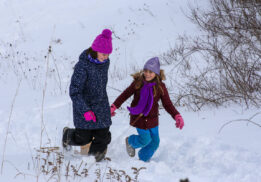 Two young girls dressed in winter clothing, including colorful hats and scarves, walking through deep snow on a snowy hillside, smiling and enjoying the cold weather.