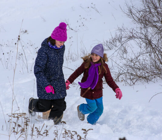 Wild Michigan at Lake St. Clair Metropark Two young girls dressed in winter clothing, including colorful hats and scarves, walking through deep snow on a snowy hillside, smiling and enjoying the cold weather.